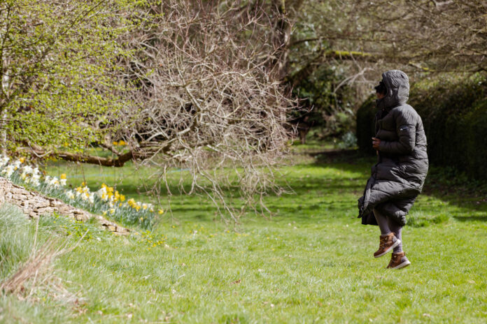 An outside scene with grass and trees. There is a women wearing a long coat with her hood up to the right hand side of the image. She is jumping off the ground.