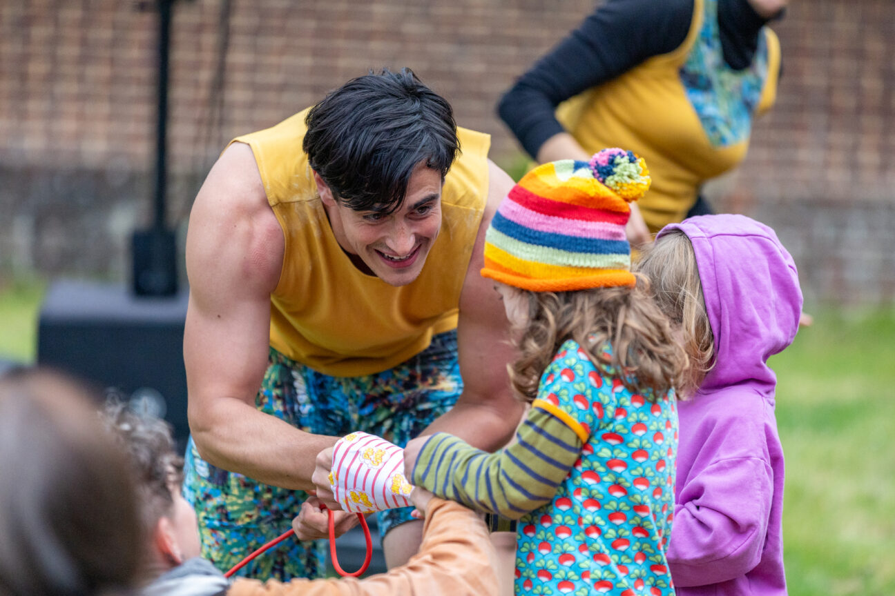 A Foley performance image with one of the performers holding a microphone unclose to some young audience members who are crowding around.