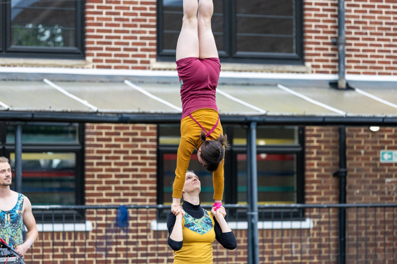 A Foley performance image showing two performers. One performer stands below holding the hands of a second performer who is doing a handstand.