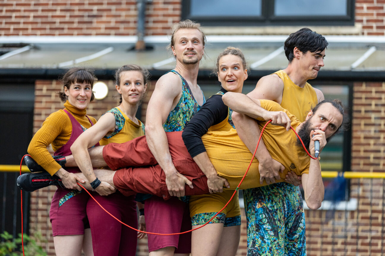 Foley performance shot including 5 performers standing in a line holding the remaining performer at a horizontal angle. The horizontal performer is holding a microphone.