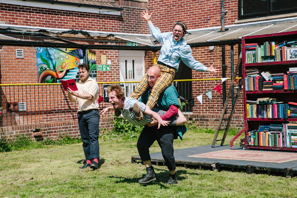 Images of Books! performance including a man standing to the left of the image reading a book whilst 3 performers create a monster like shape pulling faces at the audience