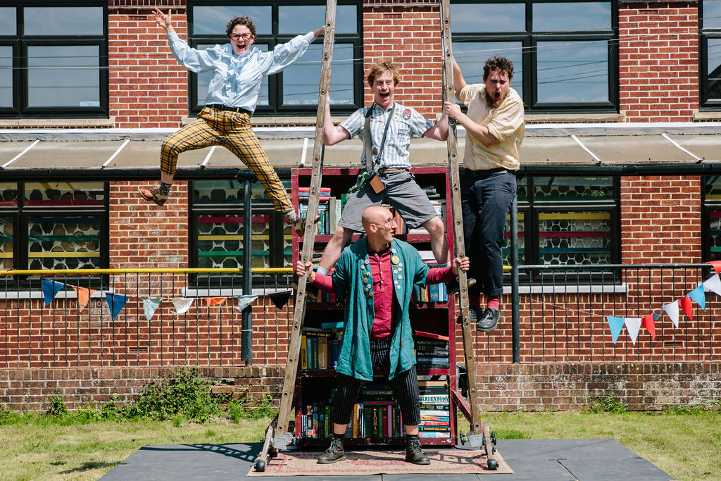 Books performance image including 4 performers in front of a bookcase. There are two ladders held up by one performer in the centre. One performer is straddling both ladders whilst the two remaining performers hang off the outside of the two ladders pulling faces at the audience